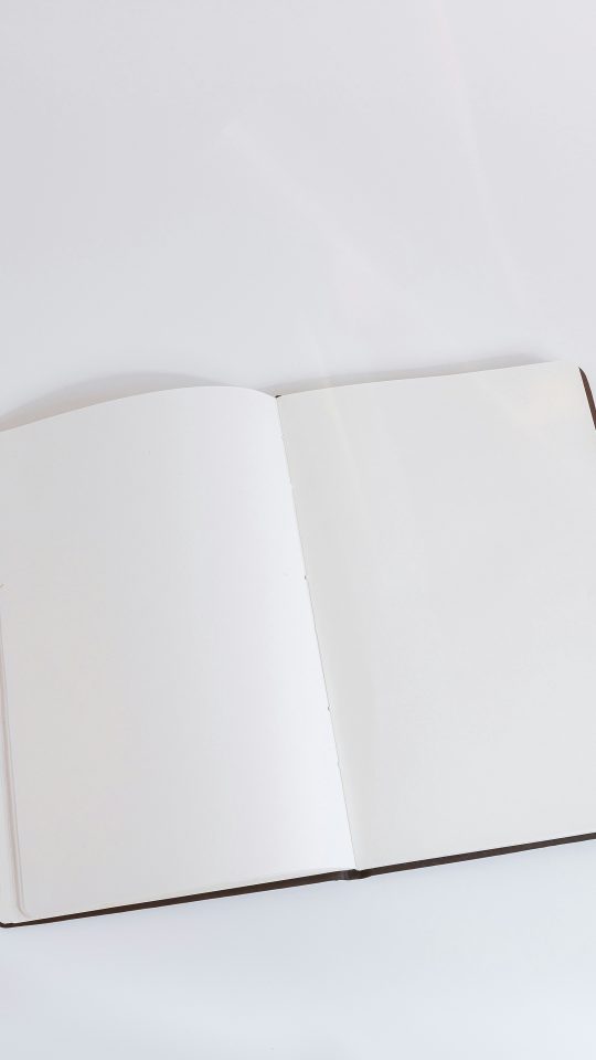 An open empty notebook on a white desk next to an iPhone and a MacBook