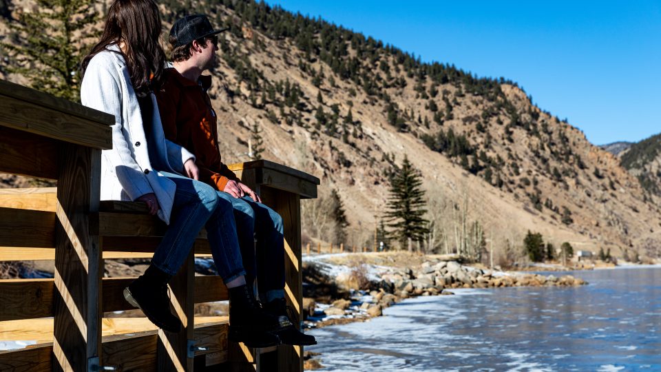 two people sitting on a wooden bench overlooking the water at The Bighorn Crossing